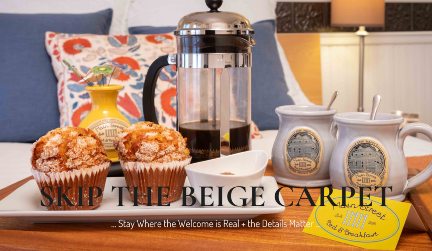A breakfast tray with fresh muffins, French press coffee, and Main Street Inn mugs set on a bed with colorful pillows, reflecting the warmth and personal details of a small boutique inn stay.