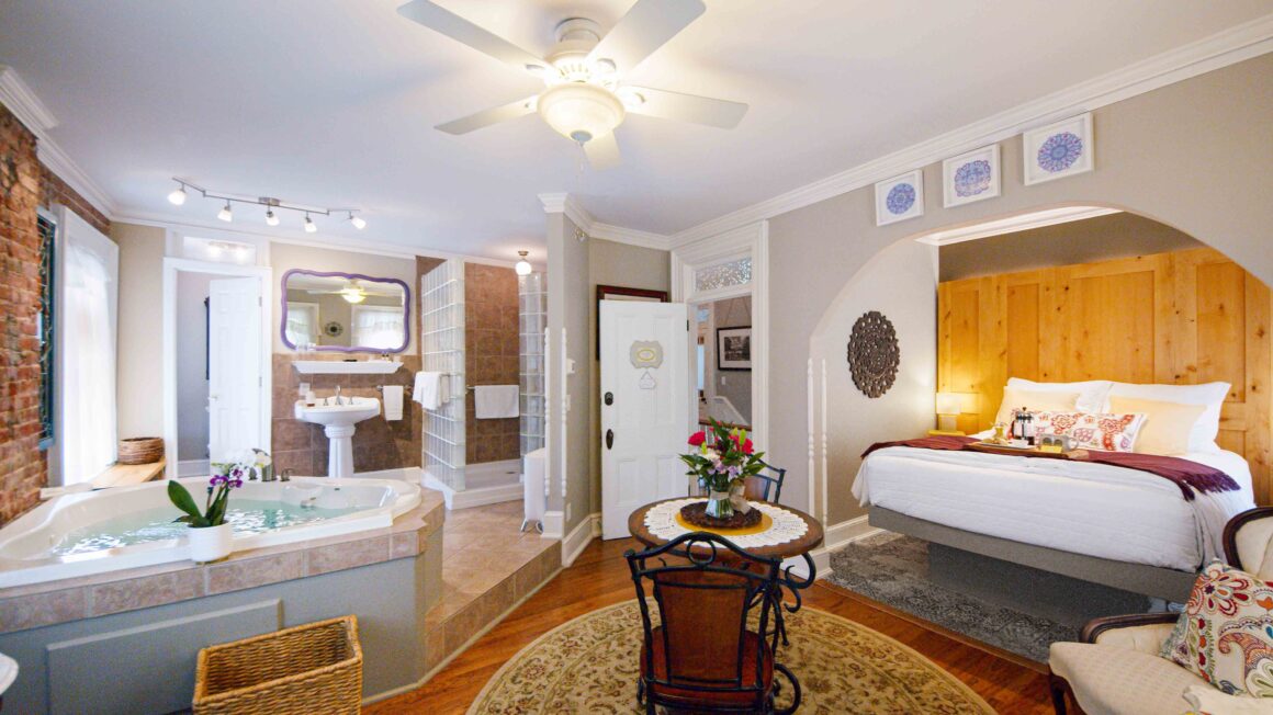 Spacious view of the Colonel Park Suite, a romantic getaway at the Main Street Inn, featuring the queen bed, sitting area, dining table, and jacuzzi tub in an elegant open layout.
