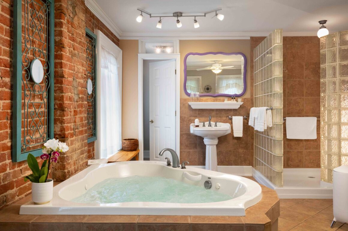 Bathroom view of the Colonel Park Suite, featuring a jacuzzi tub, pedestal sink, large mirror, and glass-block walk-in shower.