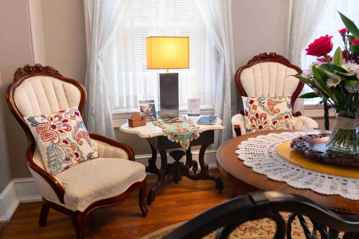 Elegant sitting area in the Colonel Park Suite featuring Victorian chairs, a small table, lamp, and lace curtains.